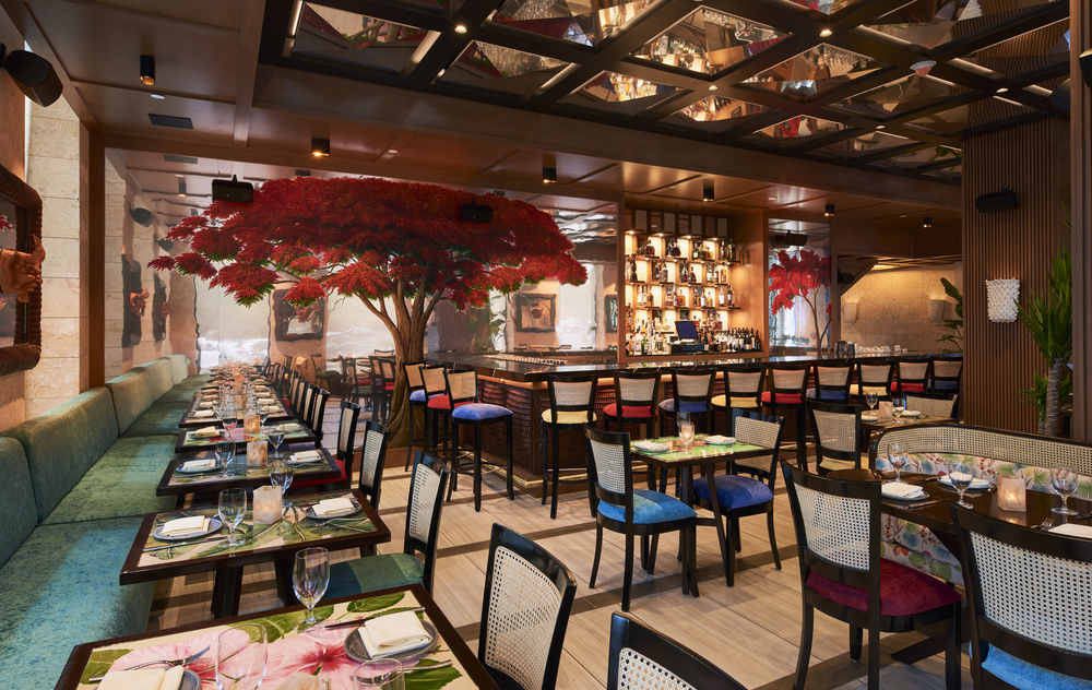 Modern restaurant with colorful chairs, neatly set tables, a central bar, and a vibrant mural of red trees on the walls, creating a lively, inviting atmosphere.