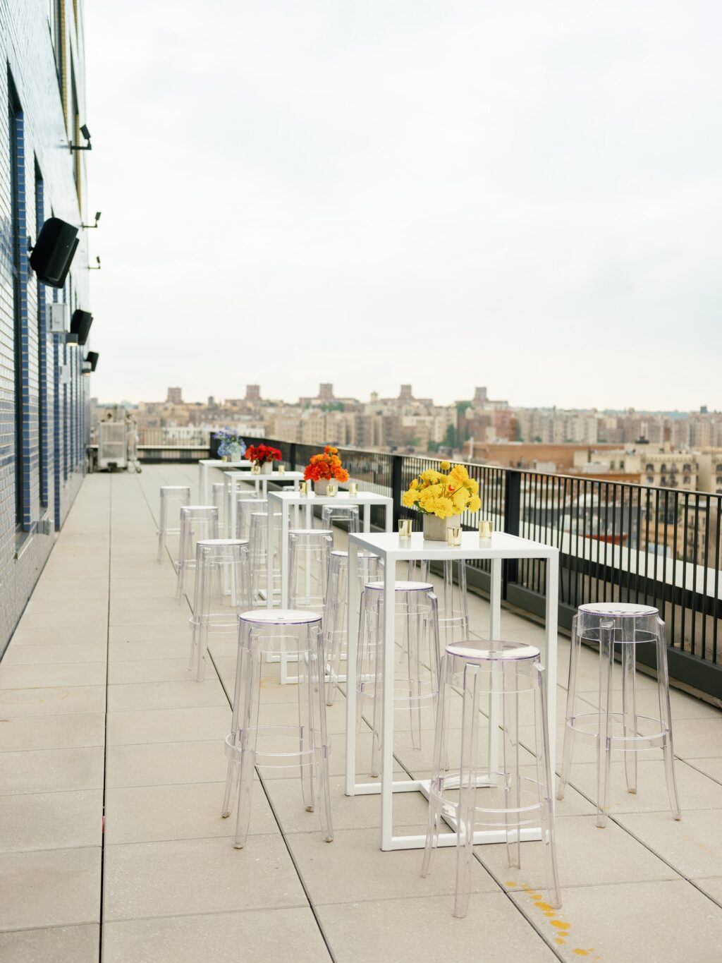 A rooftop bar with white tables, clear chairs, and flower vases overlooking the city.