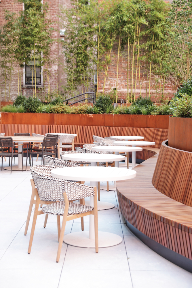 A modern outdoor seating area with wooden benches, white tables, and woven chairs.