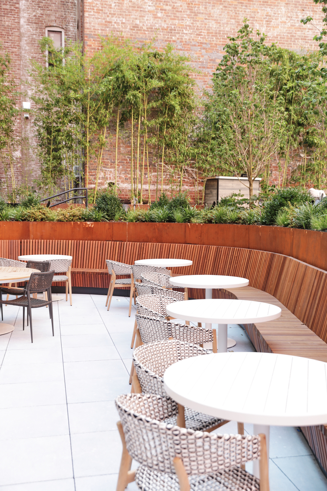 An outdoor seating area with white tables and wicker chairs, surrounded by planters and bamboo trees.