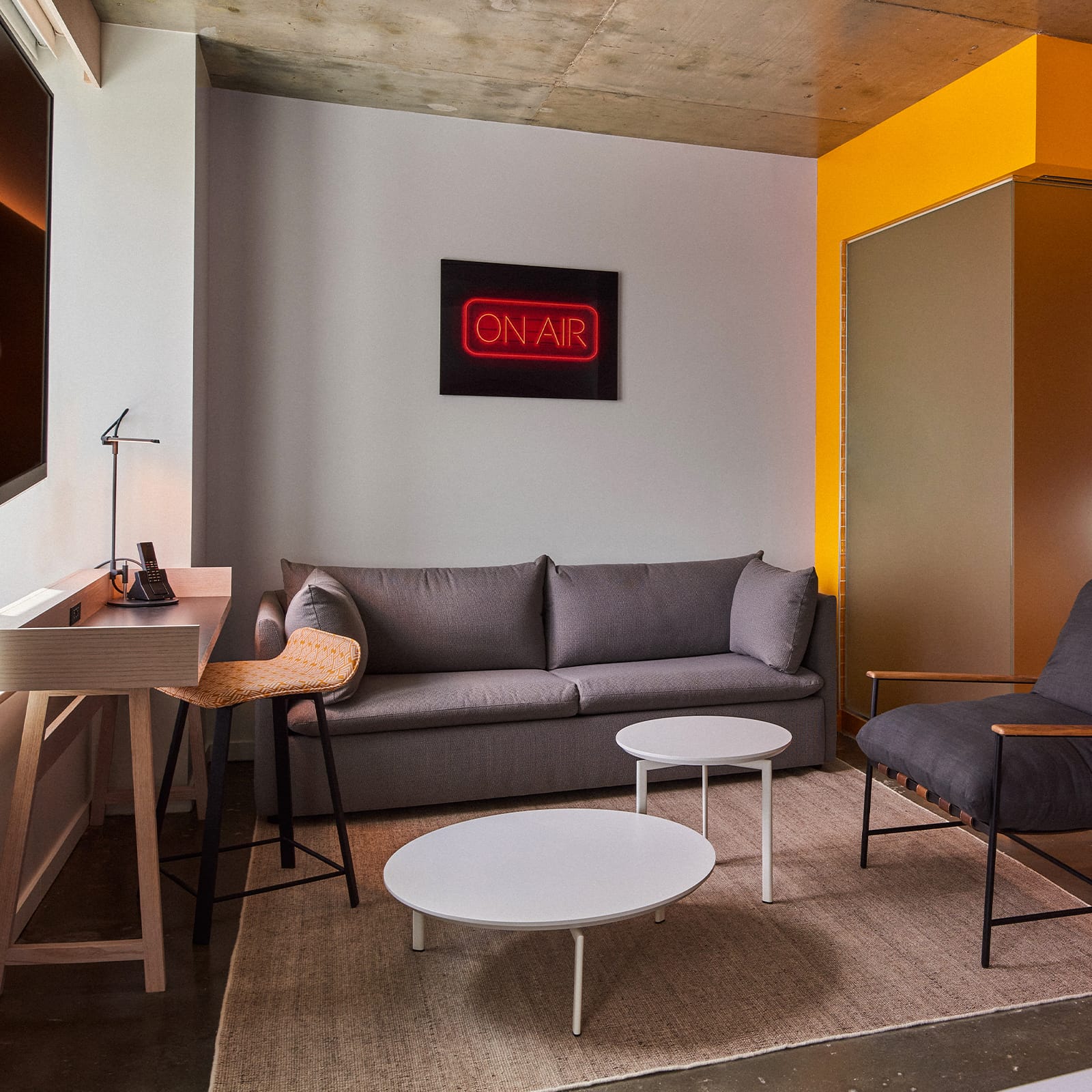 A wide view of a hotel suite with a gray sofa, two white coffee tables, a desk with a chair, and a neon 'ON AIR' sign on the wall.