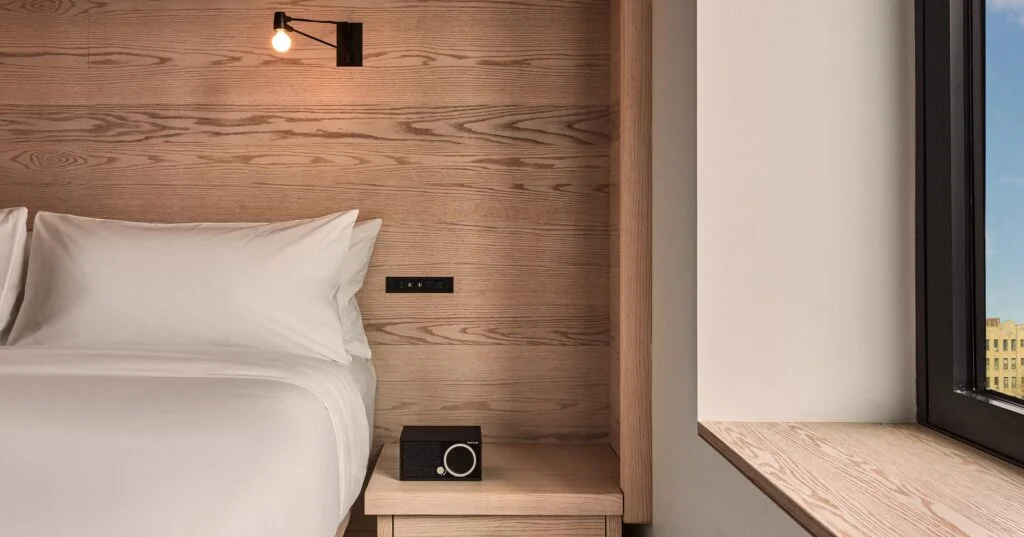 A bed with a wooden headboard, reading light, and nightstand with a radio in a room with a window.