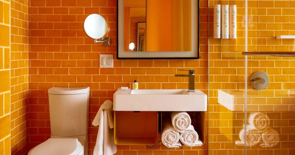 A bathroom with orange tile walls, a white sink, toilet, and mirror, with towels and toiletries.