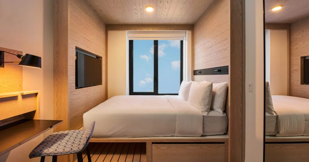 A wide view of a hotel room with a bed, window, desk, mirror, and TV.