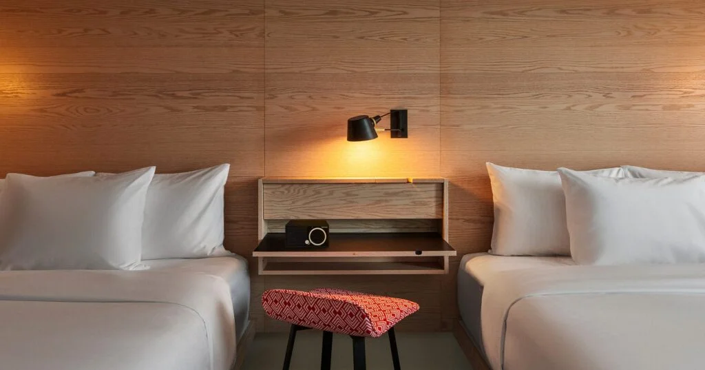 A hotel room with two double beds, wooden wall paneling, and a red ottoman.
